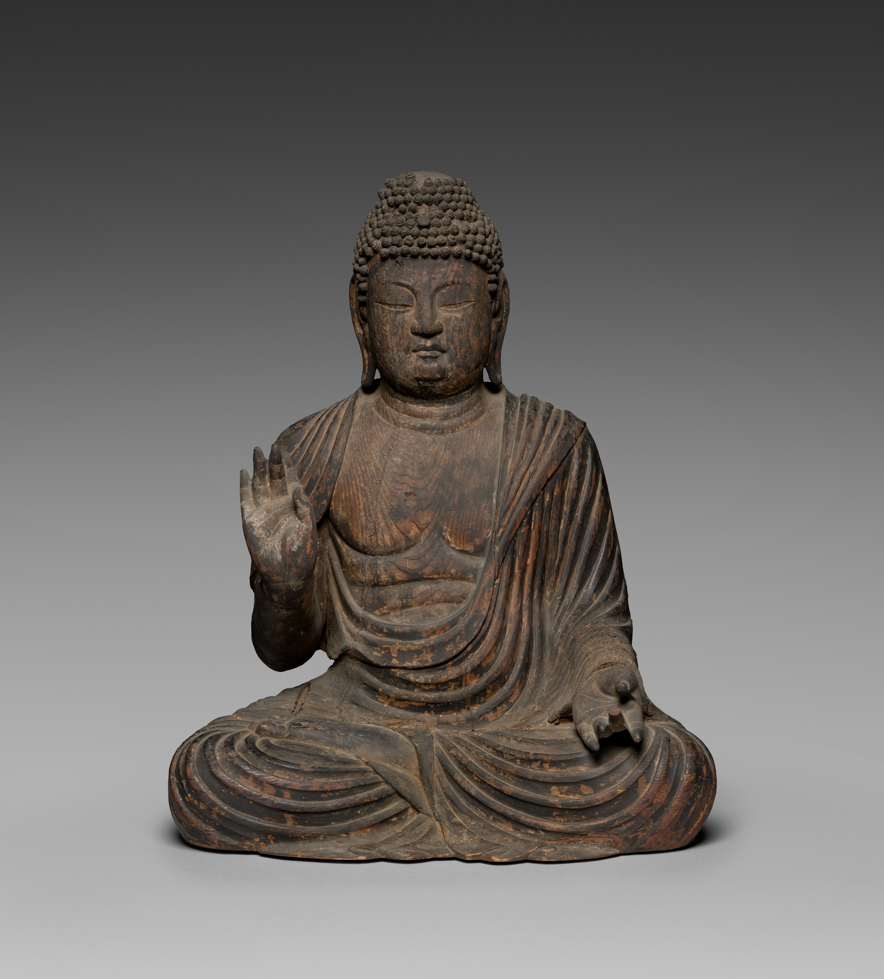 A seated wooden Buddha in cross-legged posture, right hand raised palm-out at the shoulder and left hand lowered open on the left knee, with snail-shell curls and a low cranial