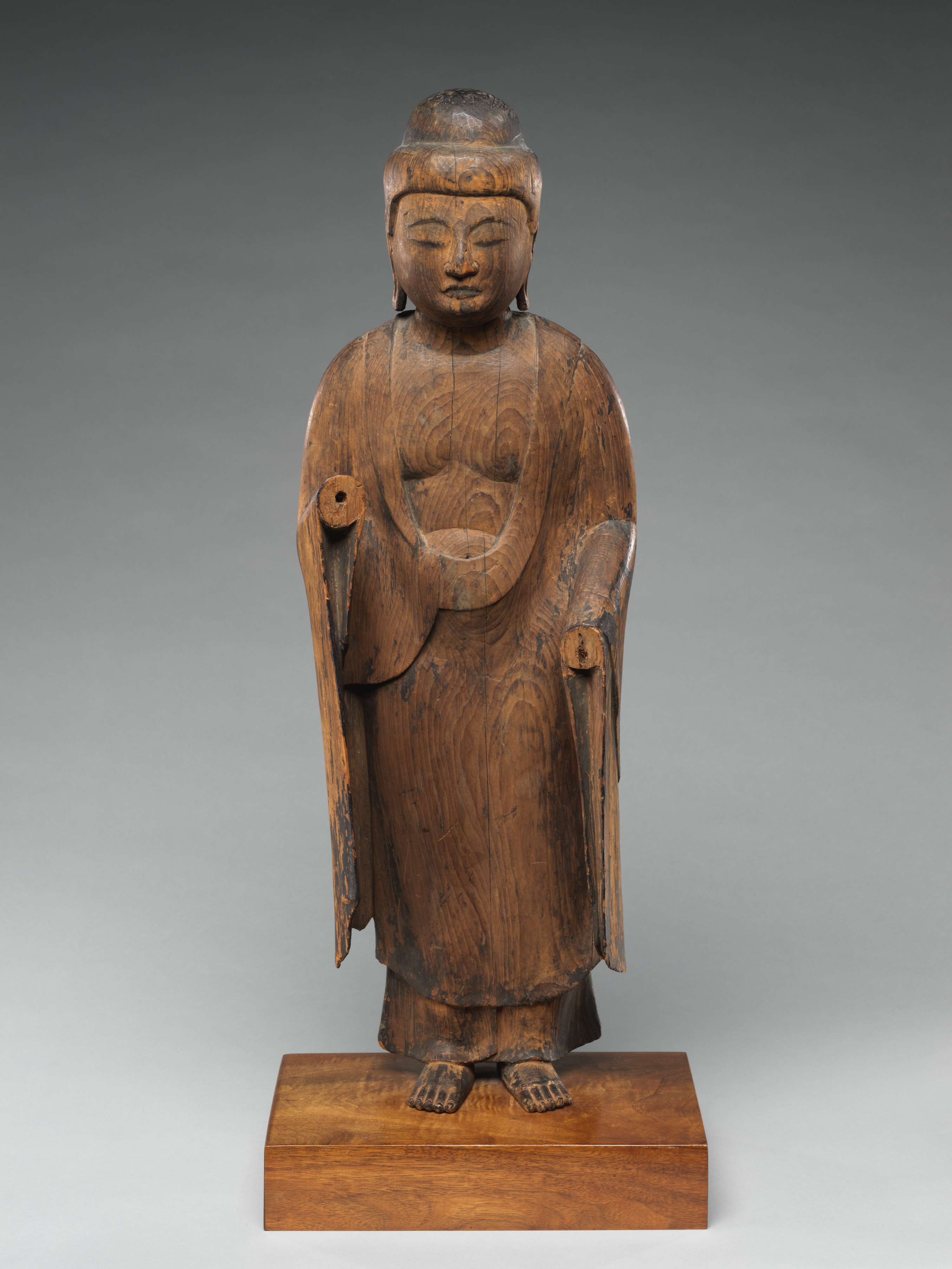 Frontal view of a small standing wooden Buddha, early Heian, the surface weathered to bare pale wood with the gilding lost and both forearms missing from the elbow, so the