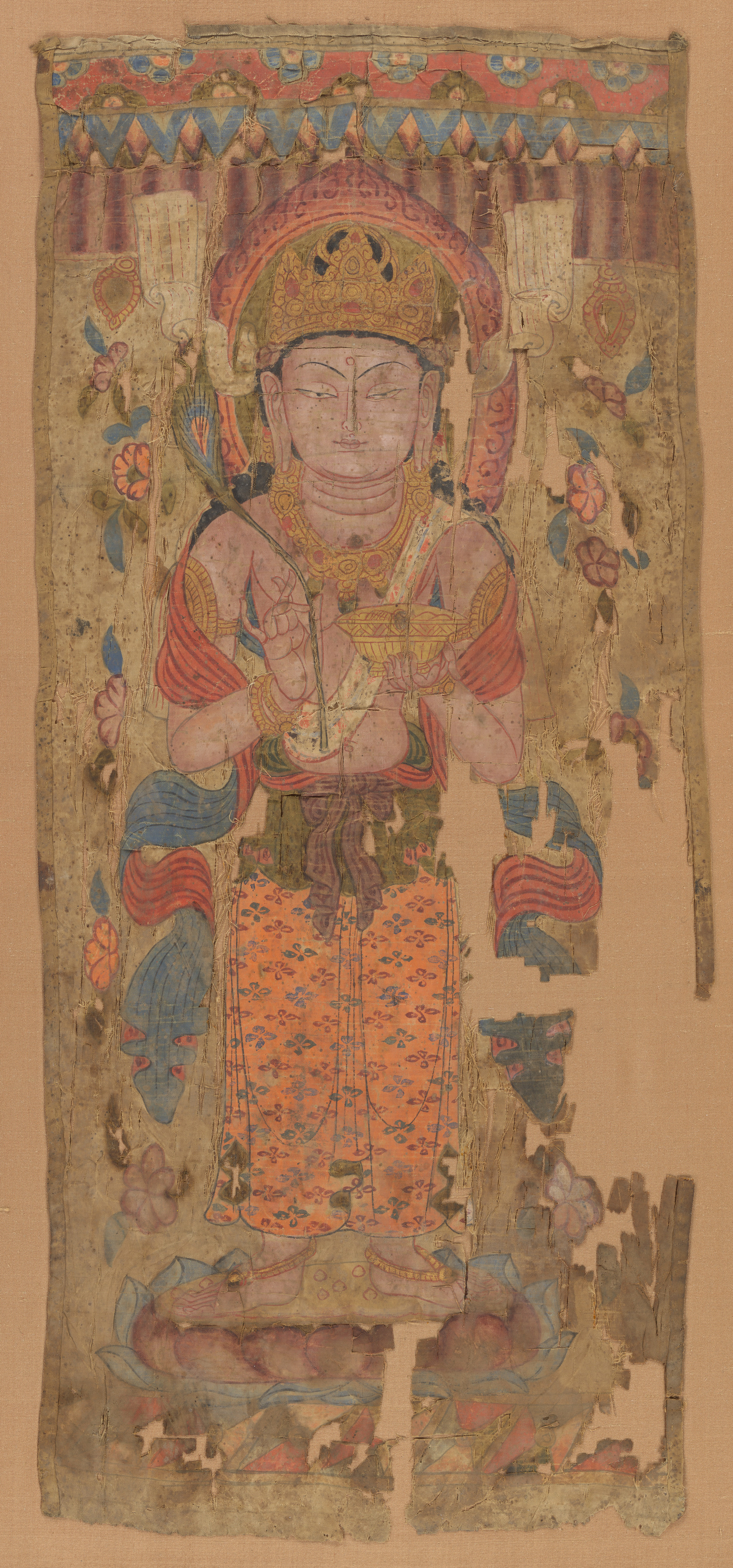 A tall narrow painted-silk banner: a standing deity on a lotus, facing front, a peacock feather in the right hand and a bowl in the left; the silk split down one side.