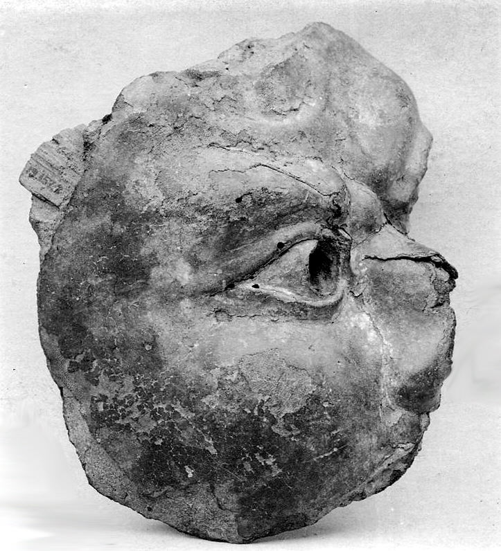 Old monochrome photograph of a small broken Buddhist head in dry lacquer: right side and crown sheared away, right eye-socket gouged open, surface granular and crumbling.