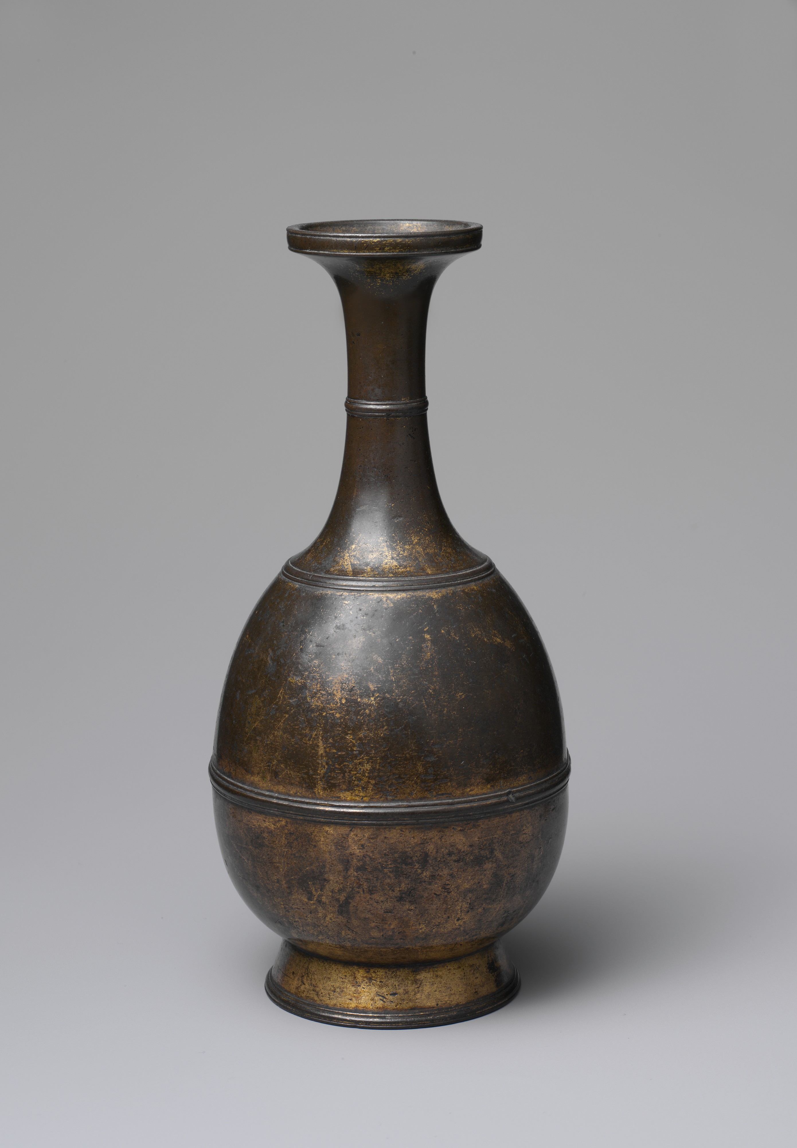 A small bottle-shaped bronze altar vessel with a flaring dish mouth, ringed neck and splayed foot; gilding survives in the hollows and is worn to dark bronze on the shoulder.