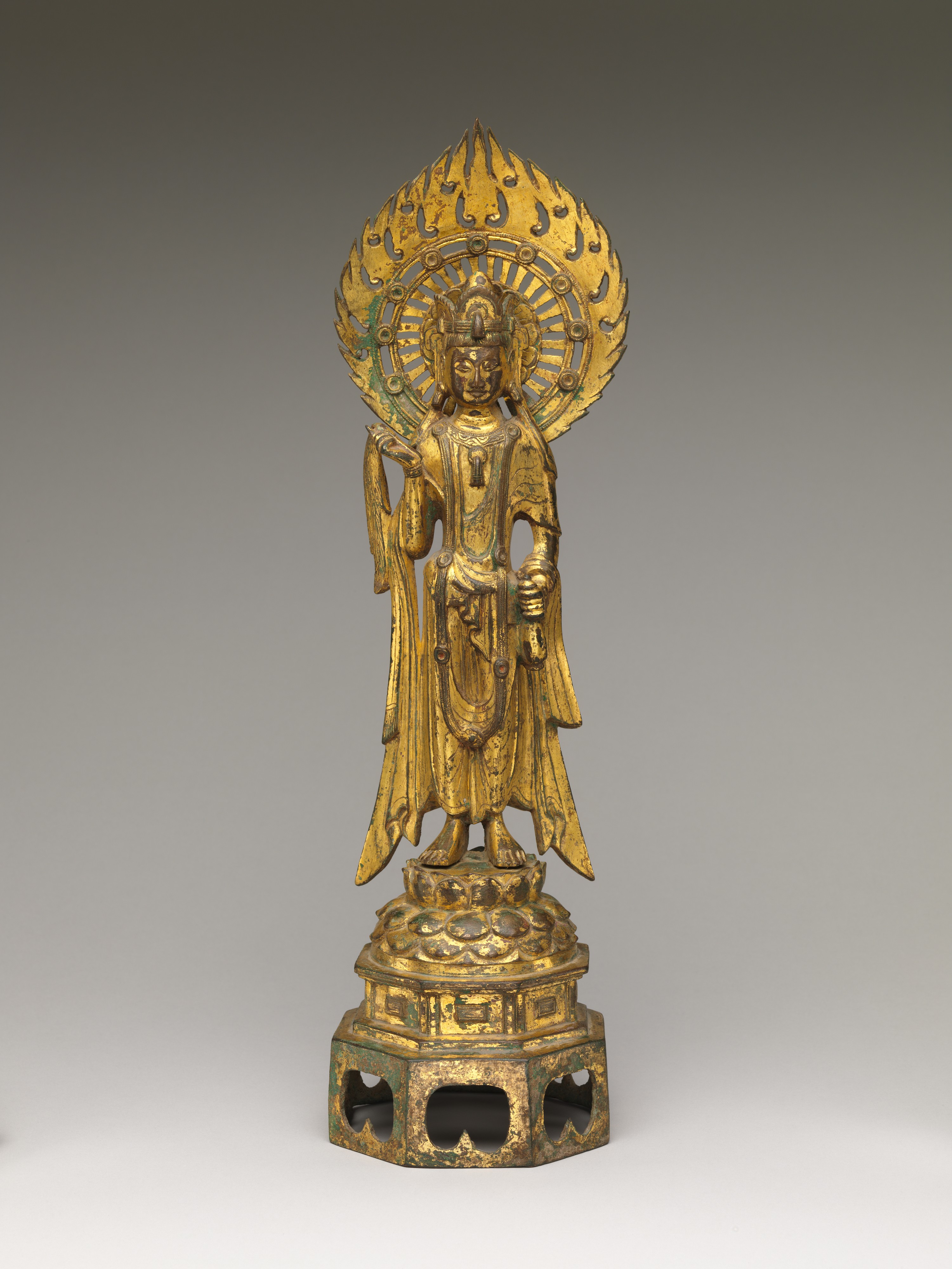A slender gilt-bronze standing bodhisattva on a lotus base, frontal and columnar, a tall crown above a long face, the left hand lifting a small vase and the right holding a willow