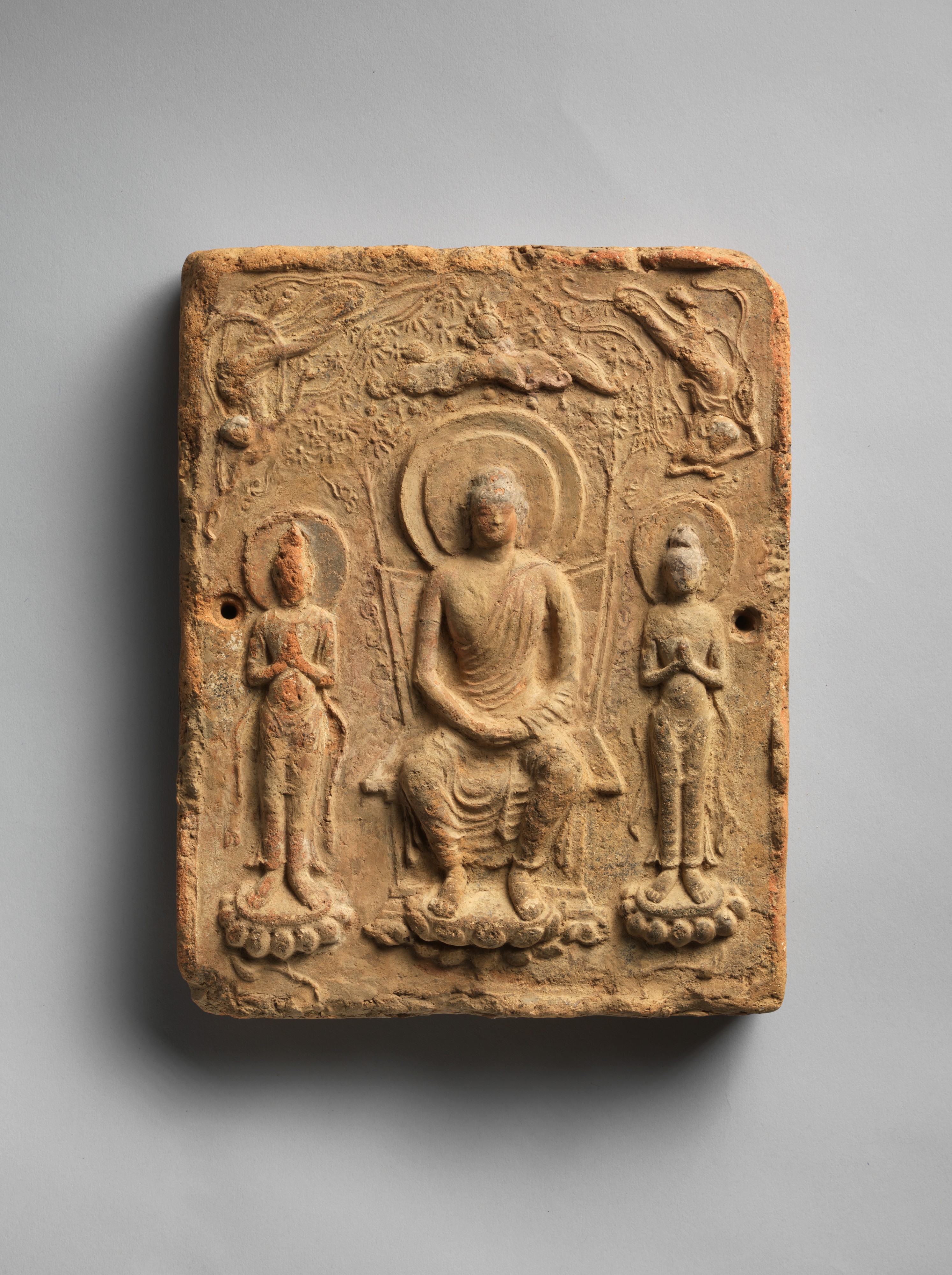 Asuka late-7th-c. earthenware relief tile, 24.5 × 19.7 × 3.8 cm. Press-moulded sanzon senbutsu: central seated Buddha beneath a canopy, two standing bodhisattvas.
