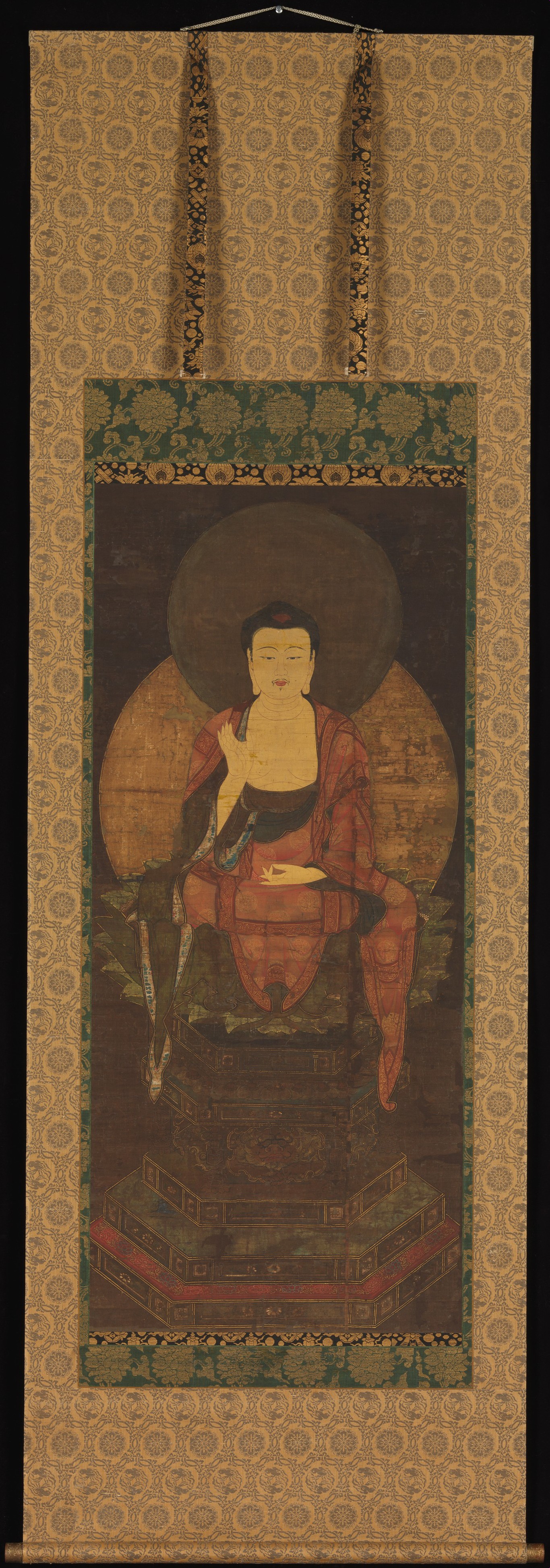 Tall hanging scroll: a gold-faced Buddha on a lotus seat above a tiered hexagonal pedestal, both legs hanging pendant, right hand raised palm-out, two mismatched halos, gold worn.