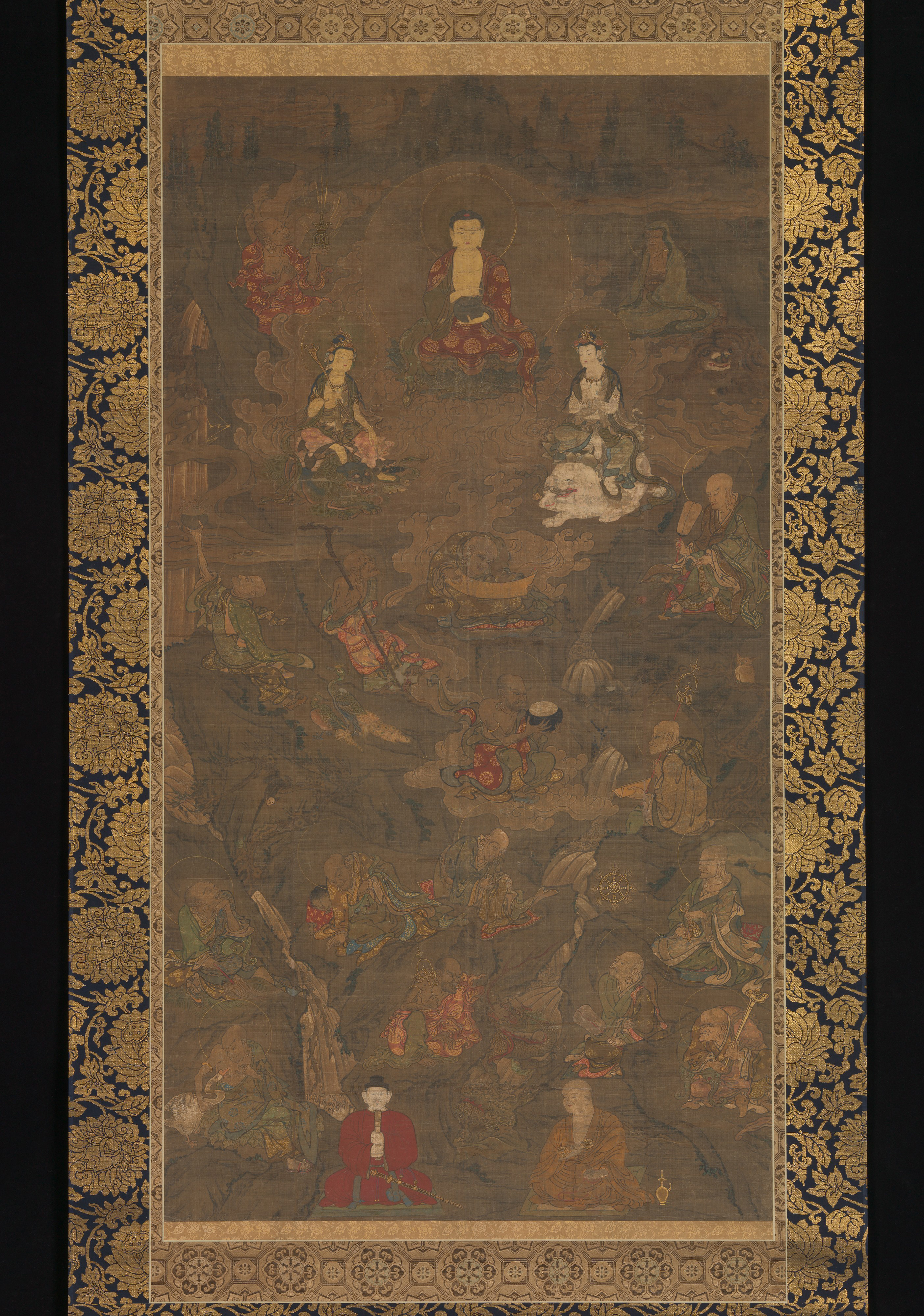 Darkened silk hanging scroll: a small gold Shaka enthroned among clouds, white bodhisattvas below, tiers of robed arhats down a rocky landscape, in a gold-brocade mount.