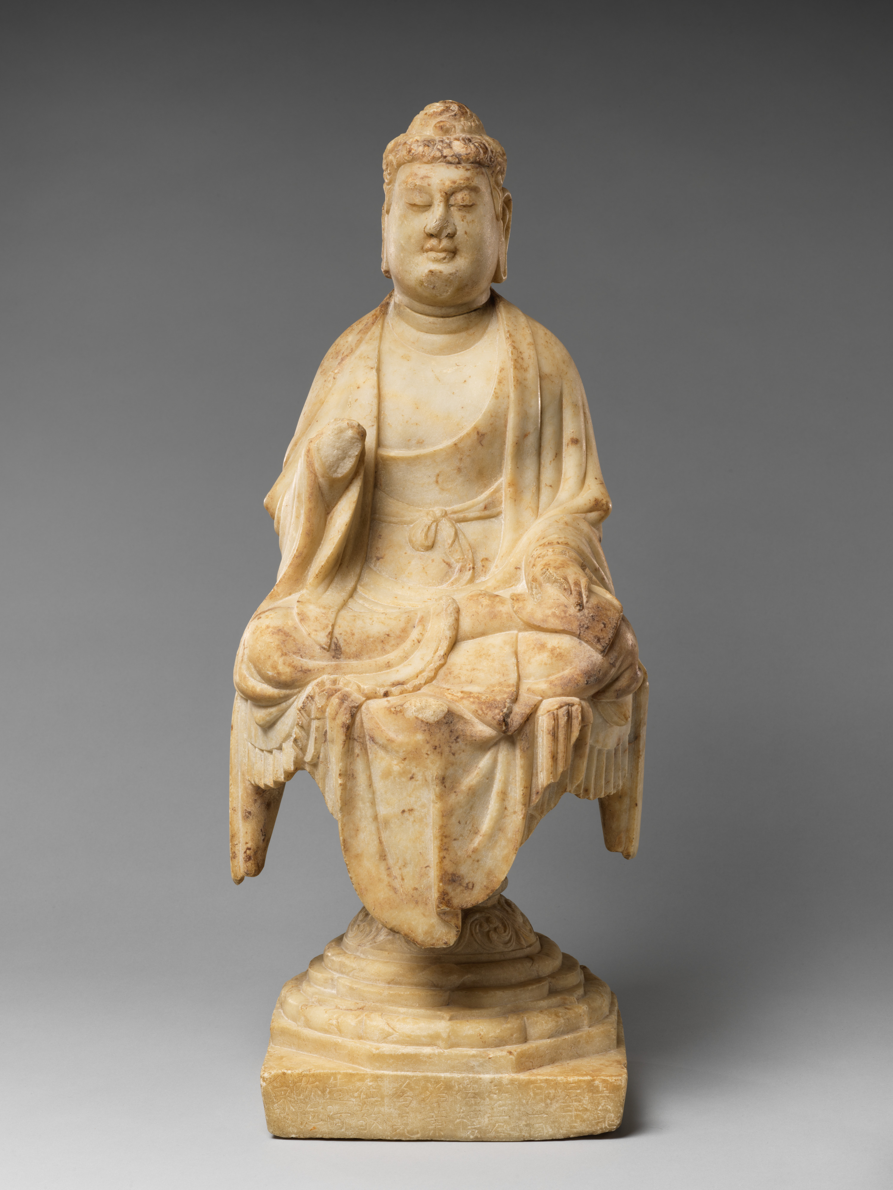 Tang Chinese marble Buddha dated 707, seated with one knee raised, drapery in heavy modelled folds over a stepped throne, rounded fleshy face, weight readable through the cloth.