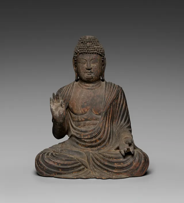 A seated wooden Buddha in cross-legged posture, right hand raised palm-out at the shoulder and left hand lowered open on the left knee, with snail-shell curls and a low cranial