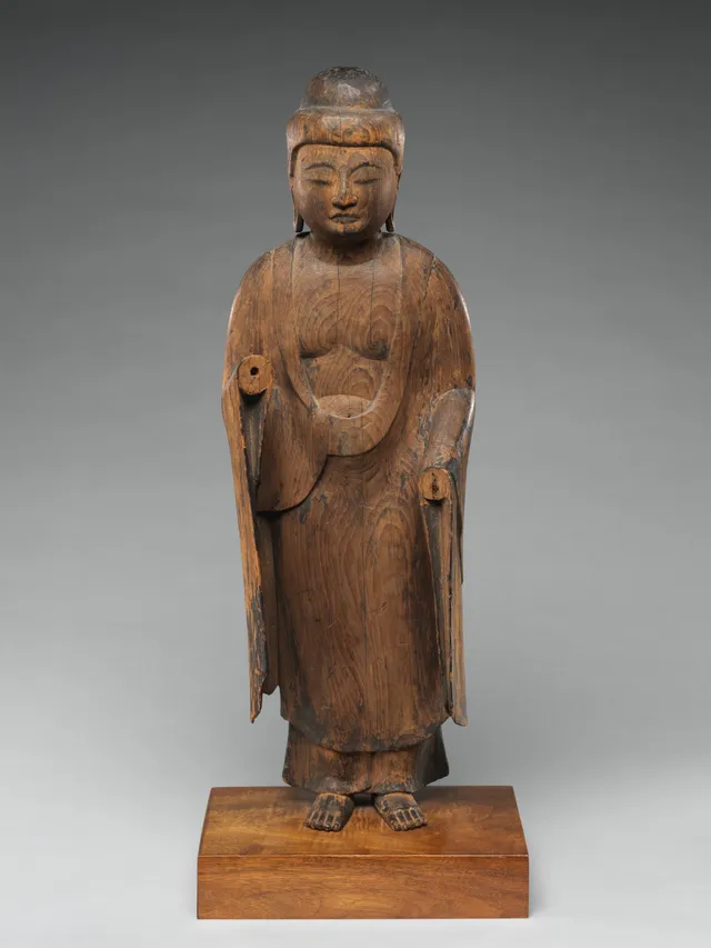 Frontal view of a small standing wooden Buddha, early Heian, the surface weathered to bare pale wood with the gilding lost and both forearms missing from the elbow, so the