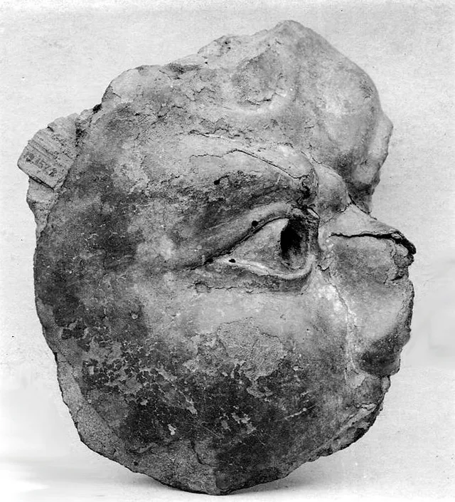 Old monochrome photograph of a small broken Buddhist head in dry lacquer: right side and crown sheared away, right eye-socket gouged open, surface granular and crumbling.