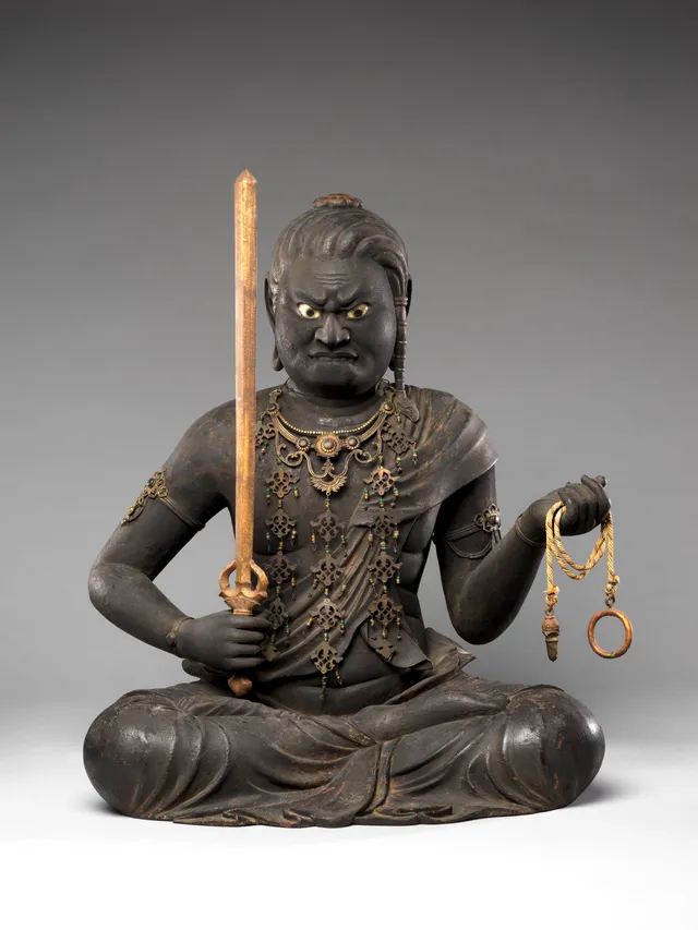 A seated wrathful wooden figure with a fierce downturned mouth and wide inset crystal eyes that catch the light, holding a sword, against a lost flame mandorla.