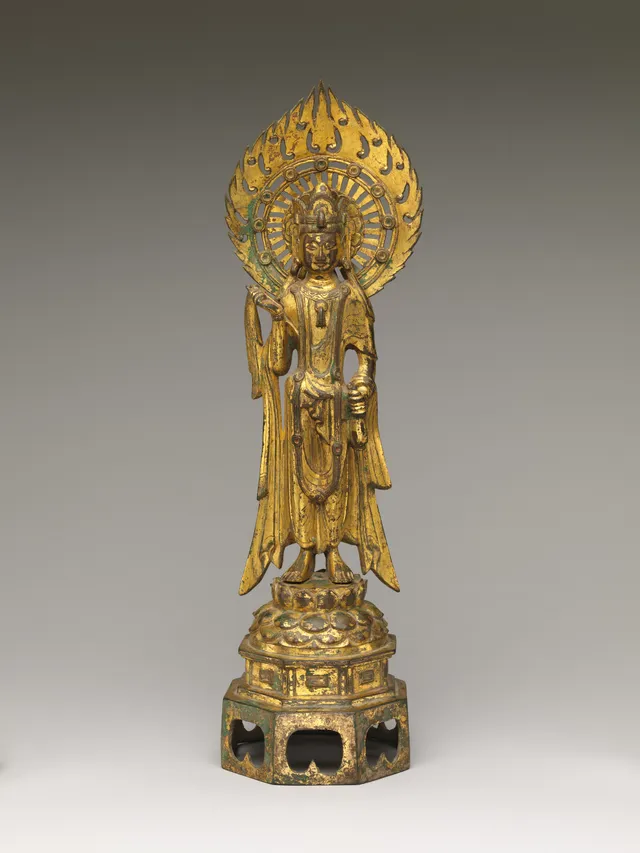 A slender gilt-bronze standing bodhisattva on a lotus base, frontal and columnar, a tall crown above a long face, the left hand lifting a small vase and the right holding a willow