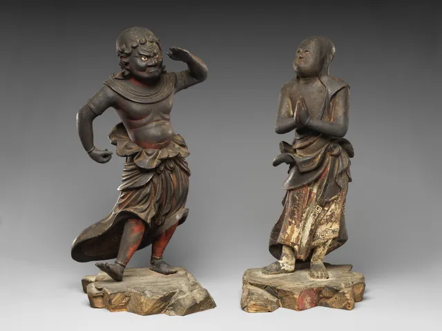 A pair of small standing wooden boy attendants on a plain ground, one with hands joined, the other holding a staff, in dark lacquered wood with worn paint and glinting inset eyes.